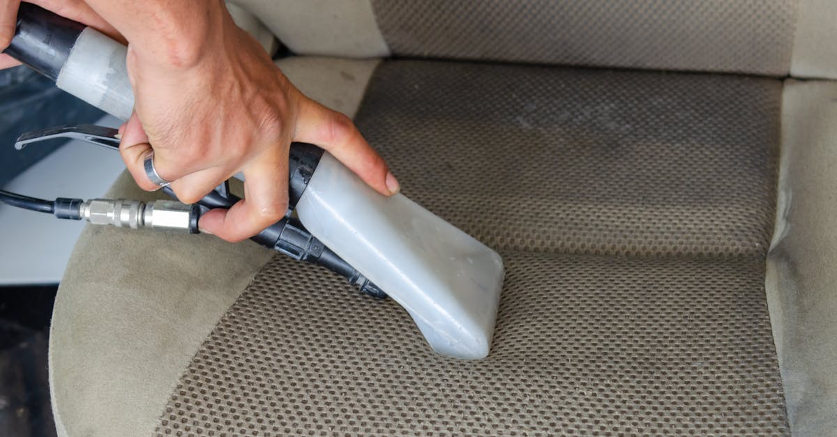 Hands using a vacuum nozzle to deeply clean a fabric car seat, removing dirt and stains.